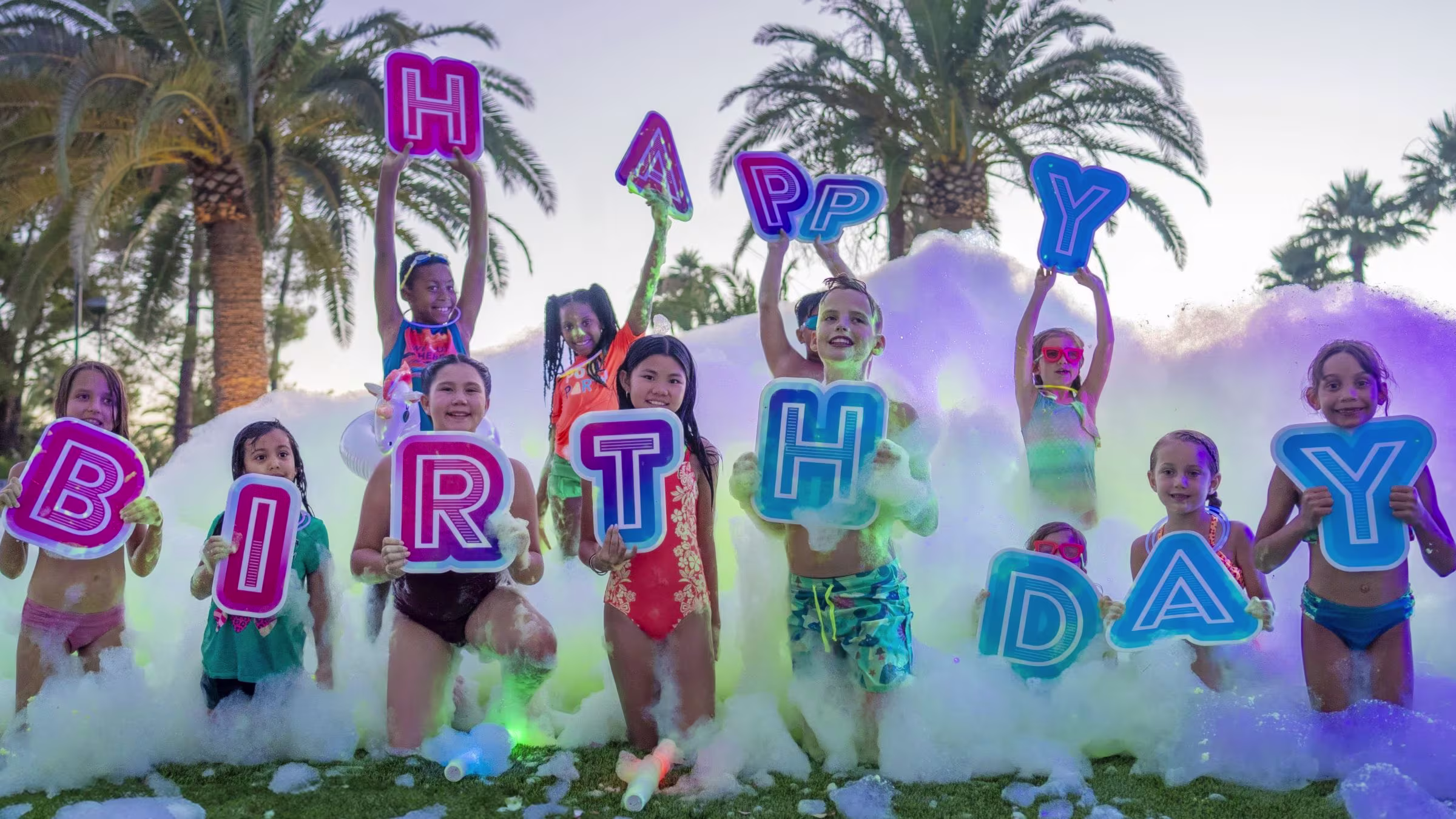 In the image, a group of six young girls are captured in a moment of joy and excitement. They are standing in a pool of white foam, their arms raised high in the air. The pool is filled with foam, creating a playful and frothy atmosphere. The girls are dressed in colorful swimsuits, adding a vibrant touch to the scene. The pool is surrounded by a fence, providing a safe boundary for the girls' fun. In the background, there are trees, adding a touch of nature to the scene.