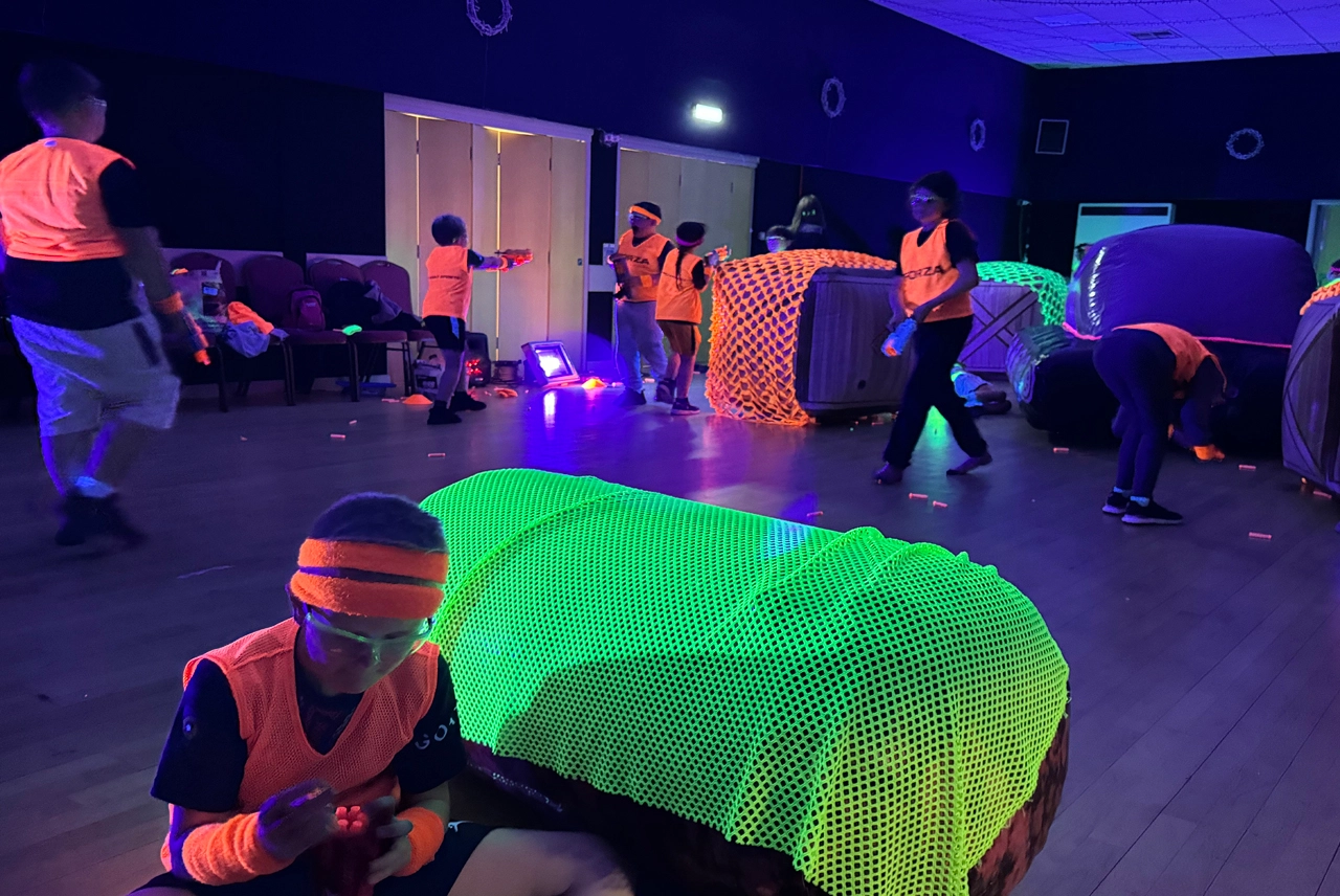 Kids playing glow Nerf battle in a dark indoor arena with glow-in-the-dark netting draped over inflatable barricades under UV blacklights.