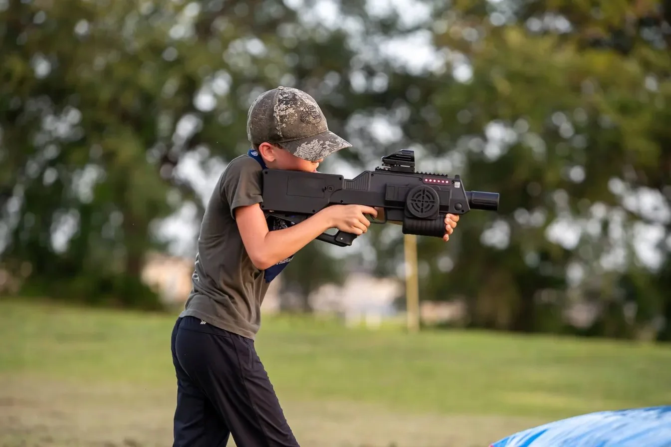 Boy aiming Halo-inspired laser tag battle rifle with red dot scope at outdoor mobile laser tag event