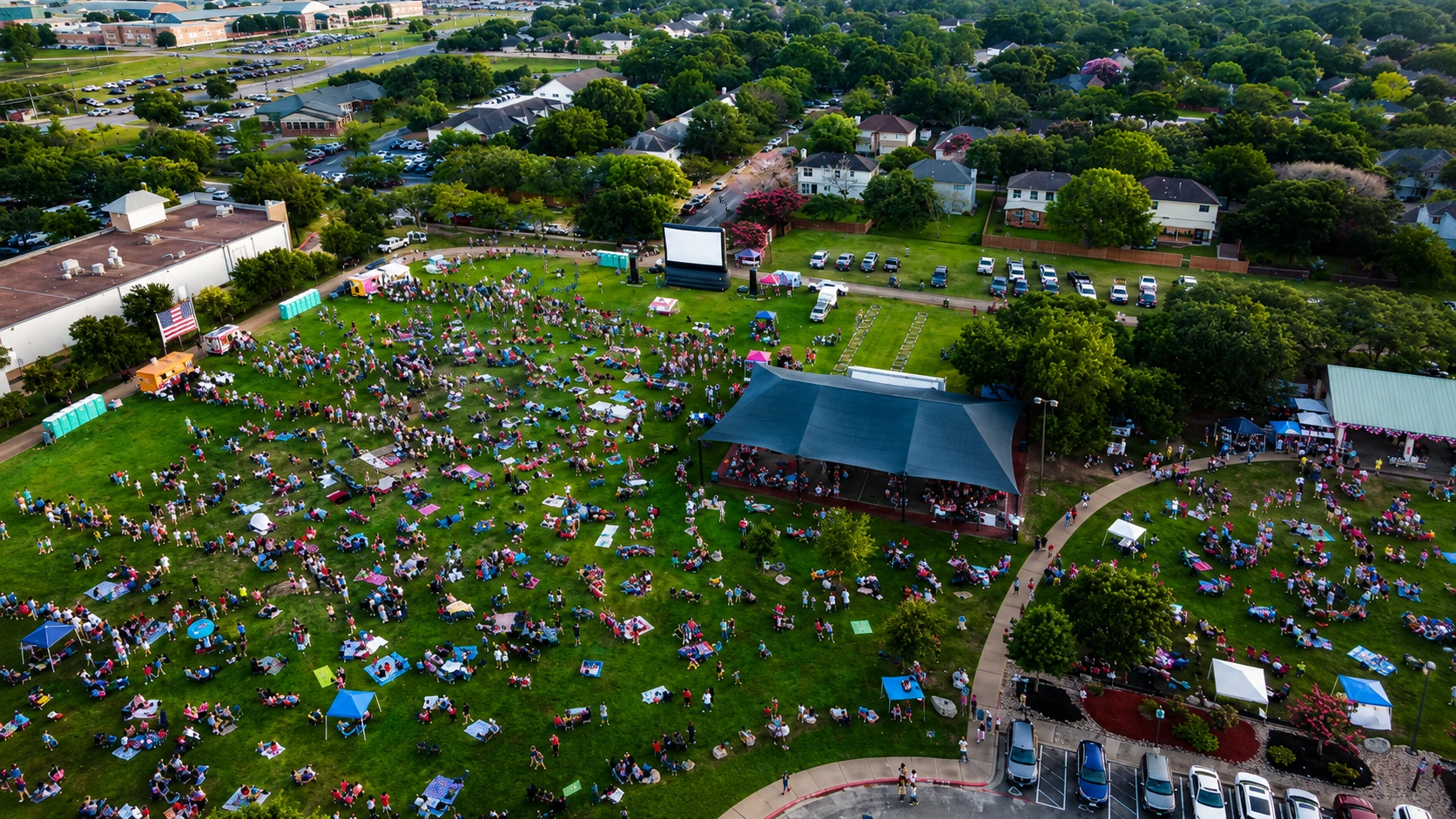 Aerial view of giant outdoor movie screen rental at large community event with thousands of guests on blankets and lawn chairs with American flag and food trucks at dusk