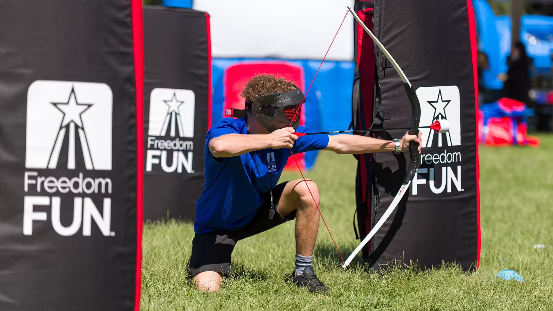 a young man is engaged in an archery game, aiming his arrow at a target. He is wearing a blue shirt and black shorts, and his face is obscured by a red mask. The target is a large black cylinder with a red star on top, and the background features a blue tent and a red banner with the word "FREEDOM FUN" written on it.