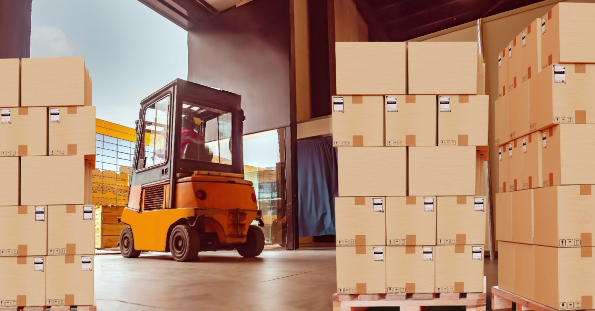 Orange forklift in a warehouse between pallets of stacked cardboard boxes.