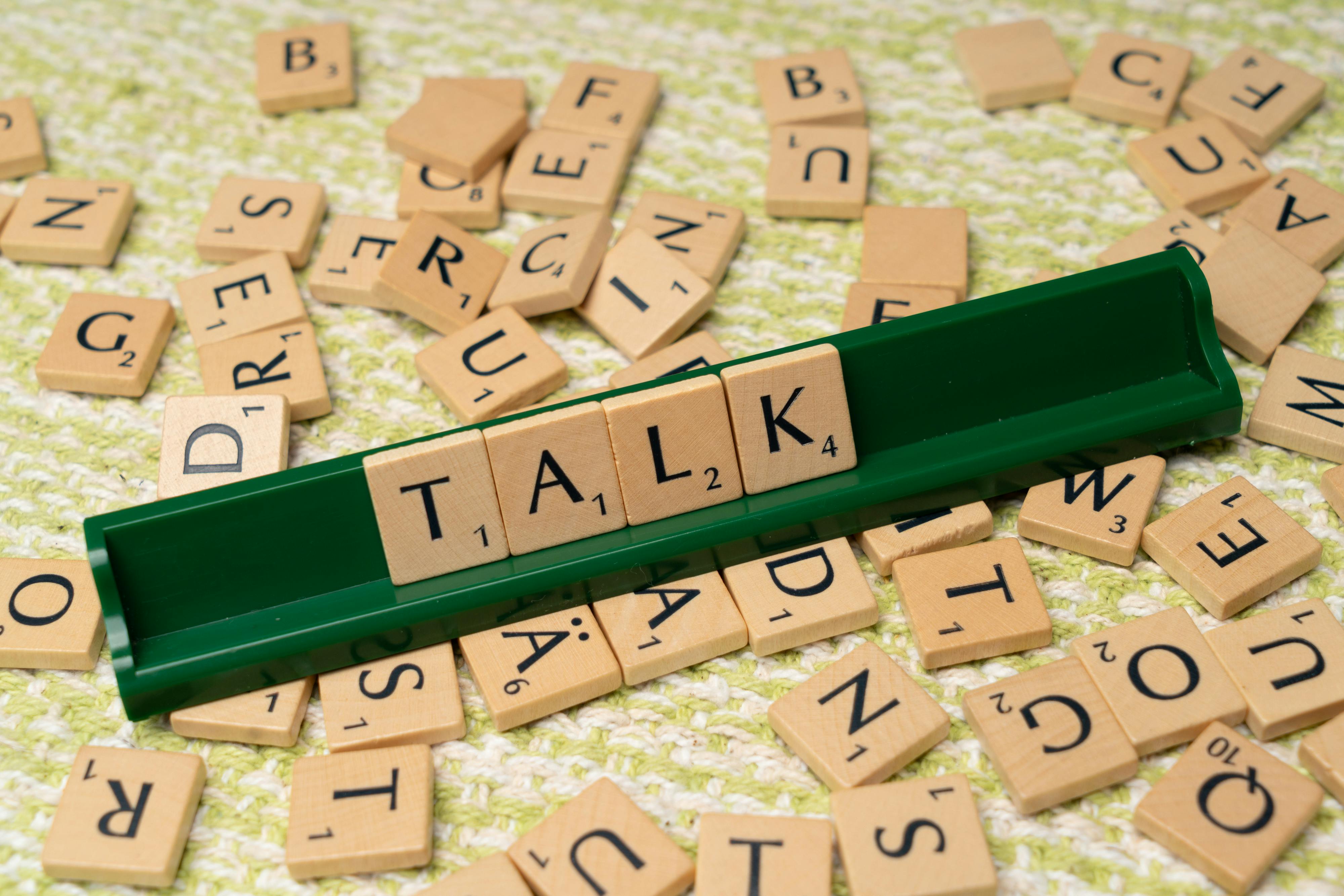 Scrabble tiles arranged on a green holder spelling the word 'TALK' surrounded by scattered letter tiles on a textured surface.