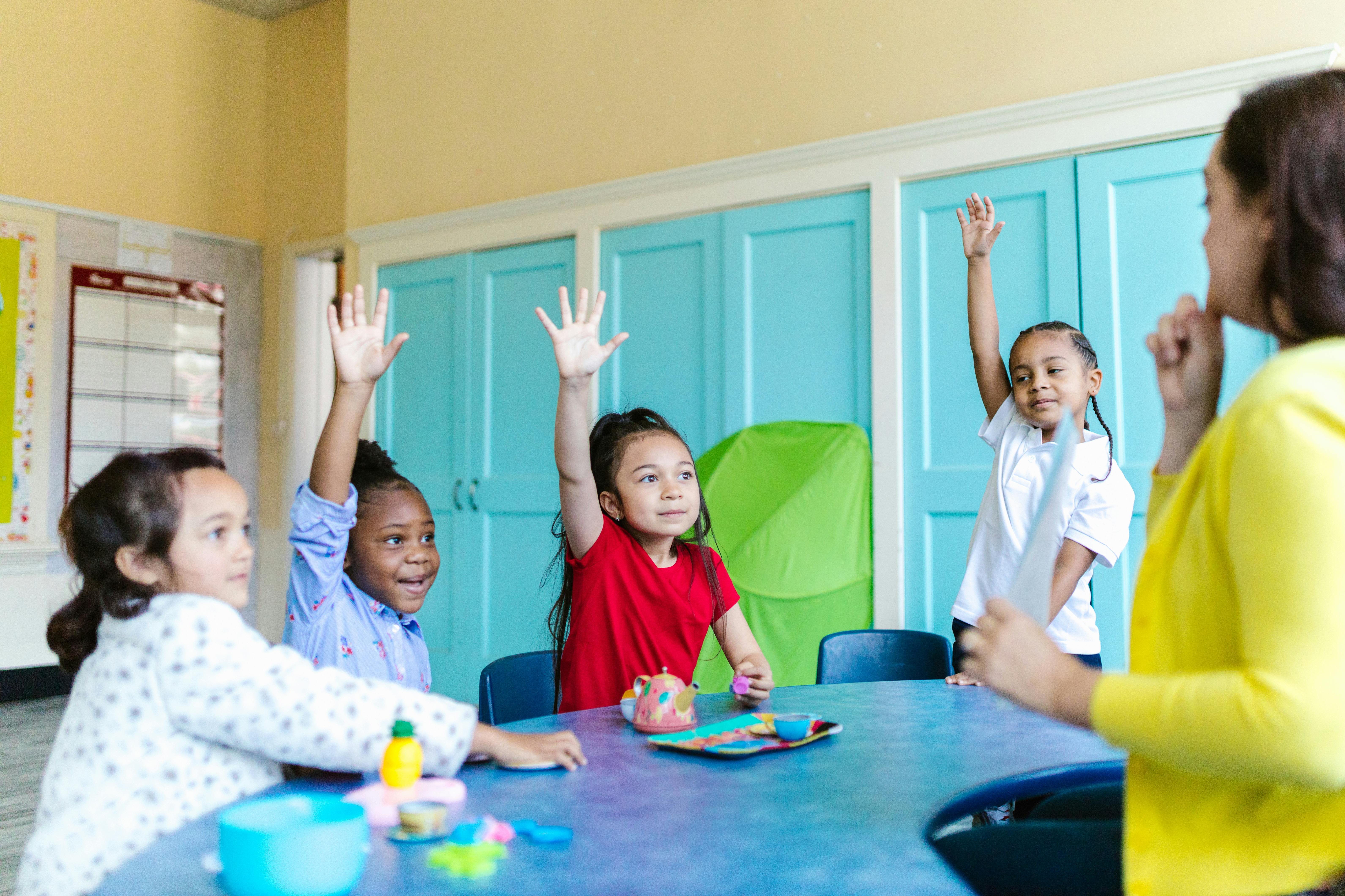 Three children at a table in a classroom raising their hands while a teacher in a yellow sweater listens.