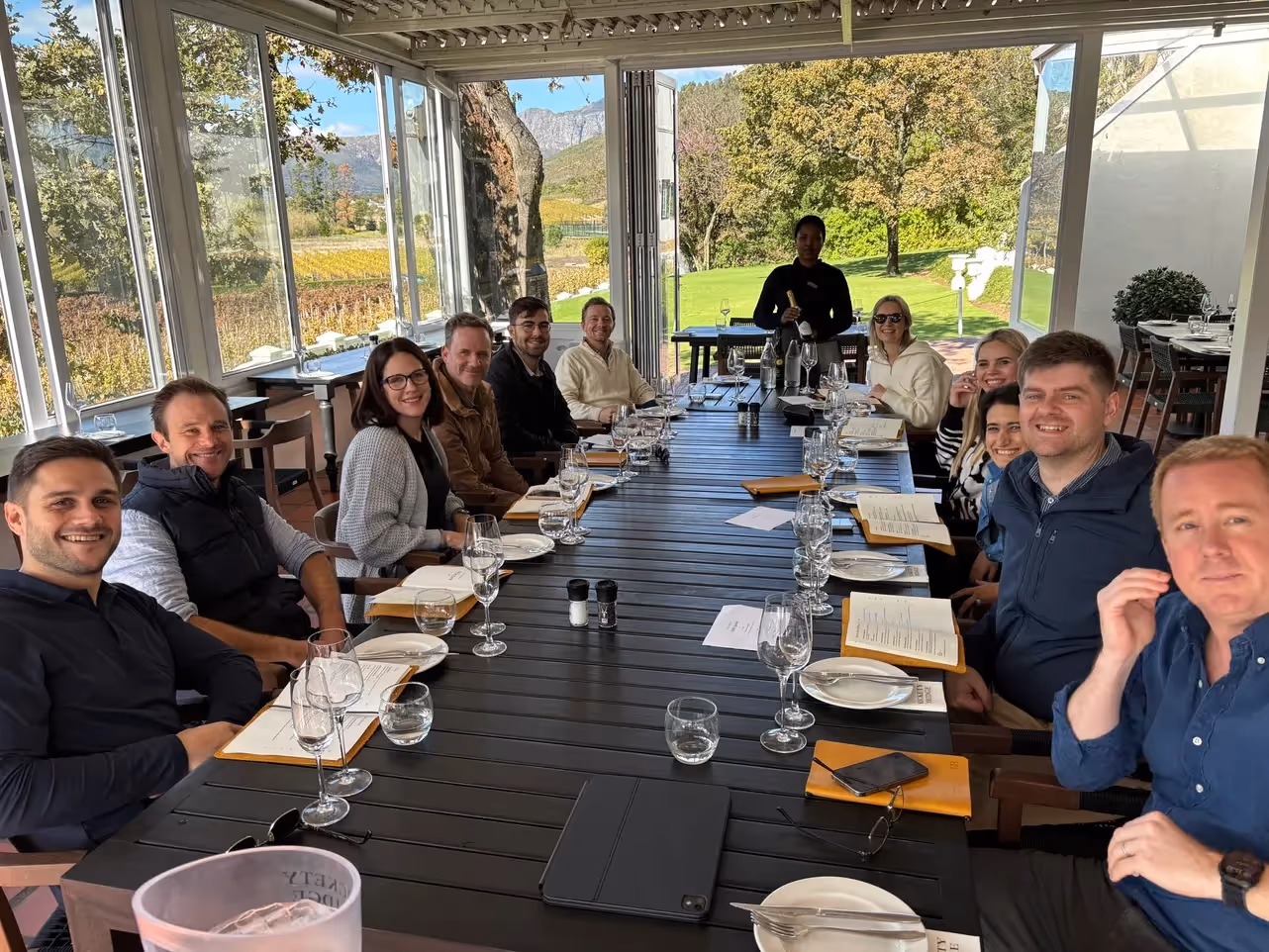 Group of twelve people seated around a long table in a bright dining room with large windows showing a scenic outdoor view, and a server standing at the head of the table.