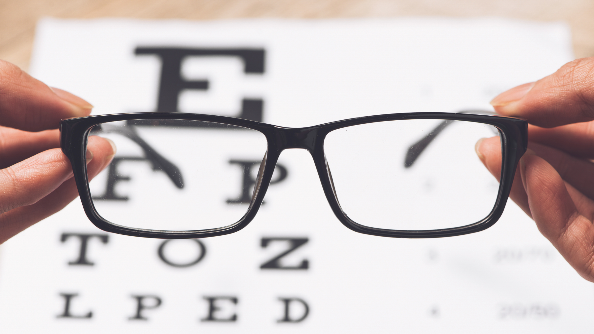 Hands holding up eye glasses frame in front of Snellen chart 