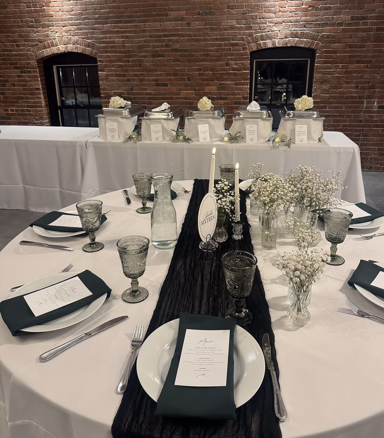 Round dining table set with white tablecloth, black table runner, white plates with dark green napkins, decorative glassware, candles, and baby's breath flowers, with buffet chafing dishes in front of a brick wall.