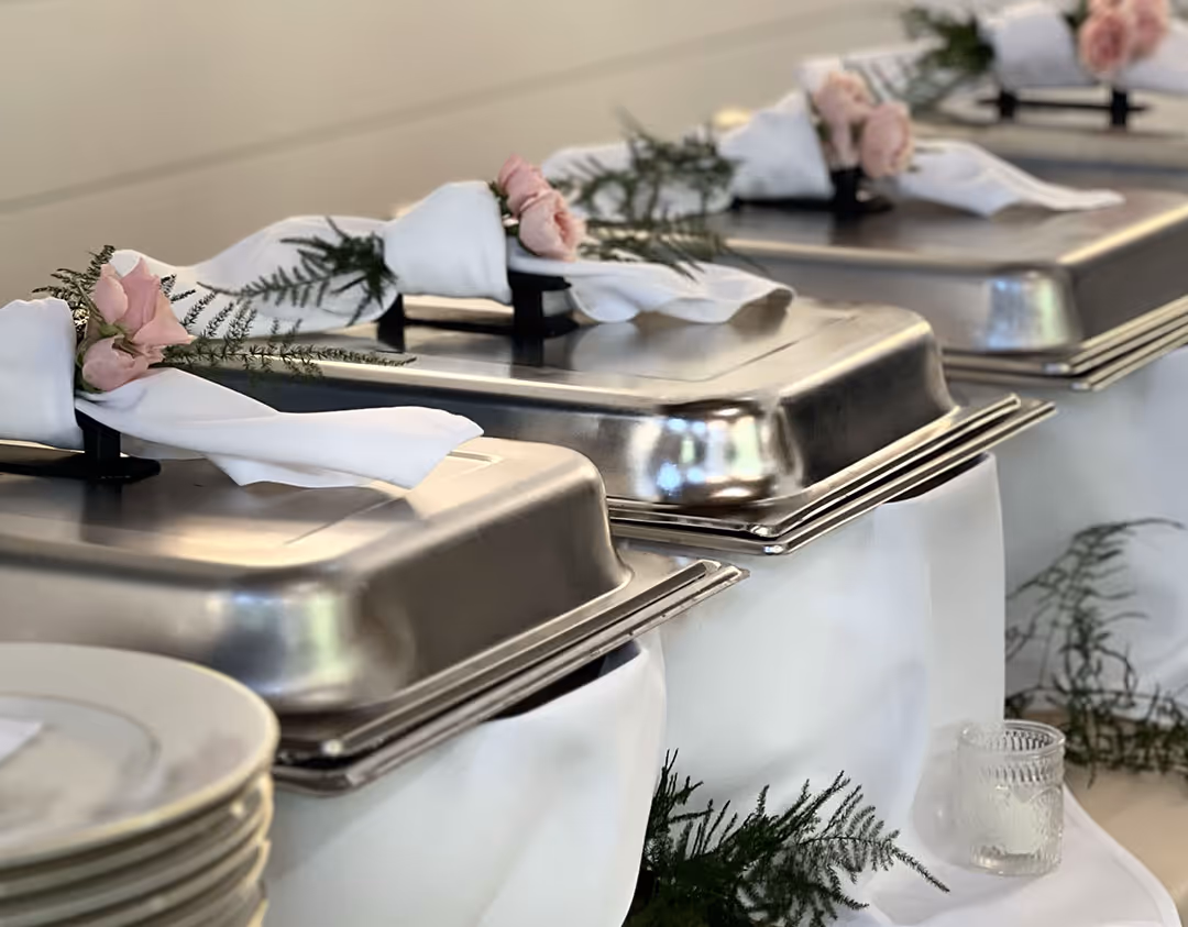 Close-up of metal buffet chafing dishes decorated with white napkins, pink roses, and green foliage on a white tablecloth.