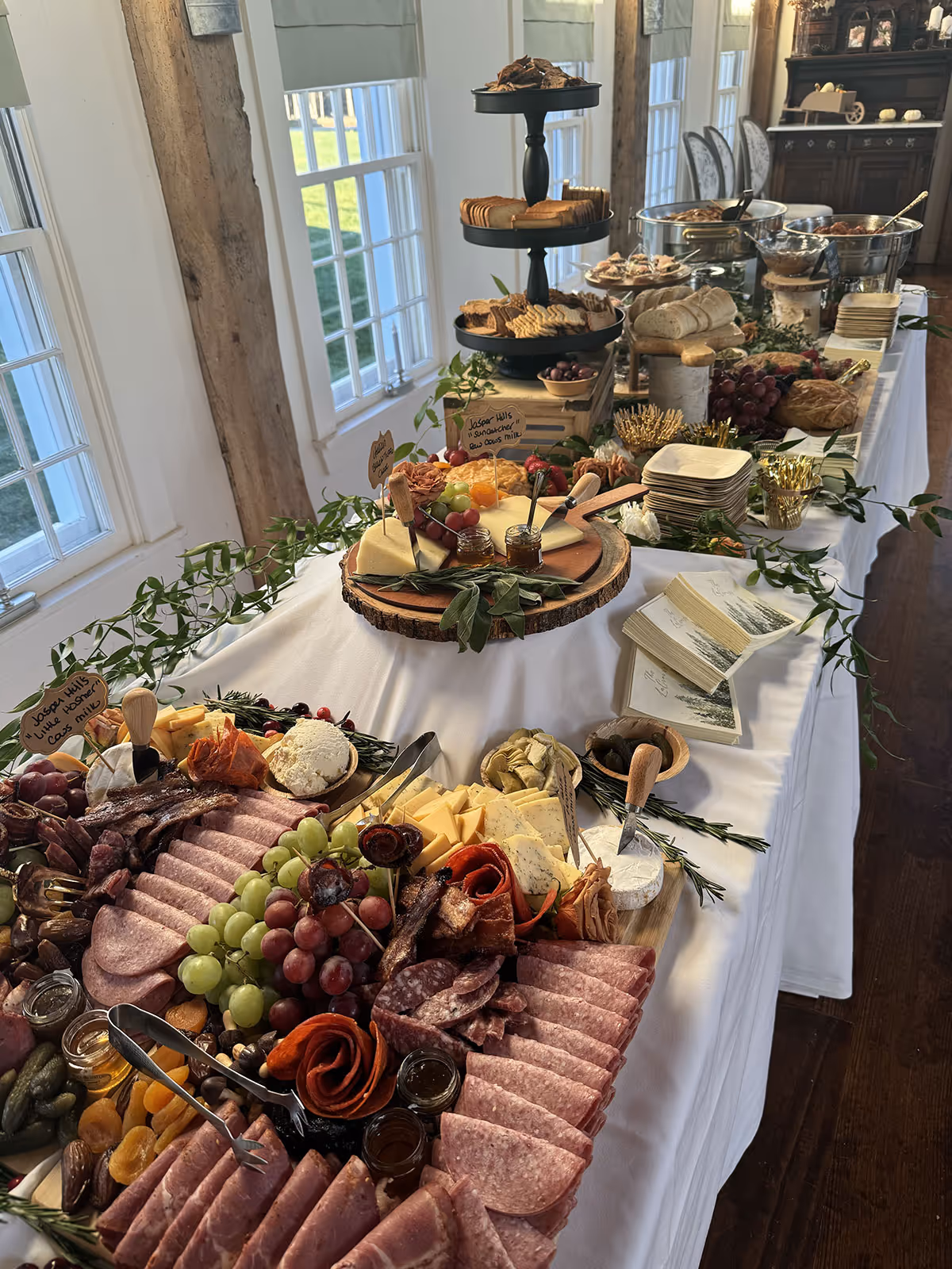 Long buffet table with a variety of charcuterie items including sliced meats, cheeses, grapes, crackers, bread, and preserves, decorated with greenery near large windows.