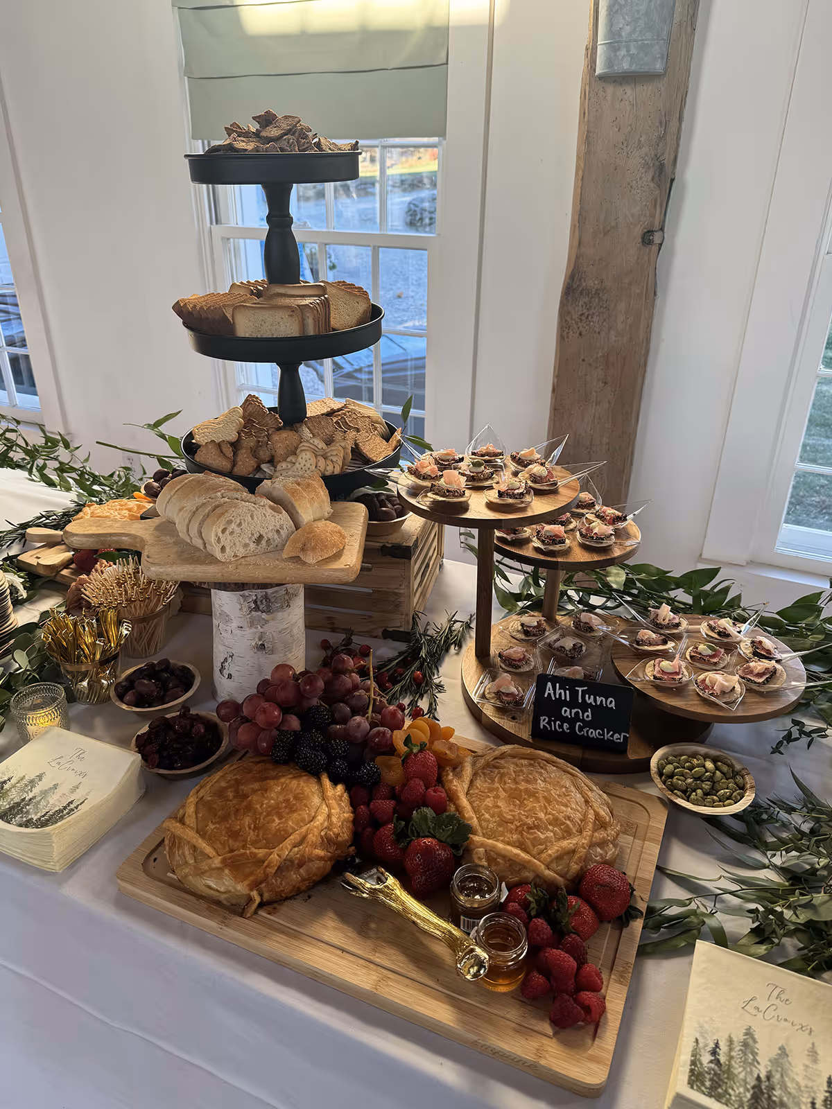 Table with a variety of appetizers including bread slices, crackers, two pies on a wooden board with fresh berries, and small servings of ahi tuna on rice crackers displayed on a tiered stand.