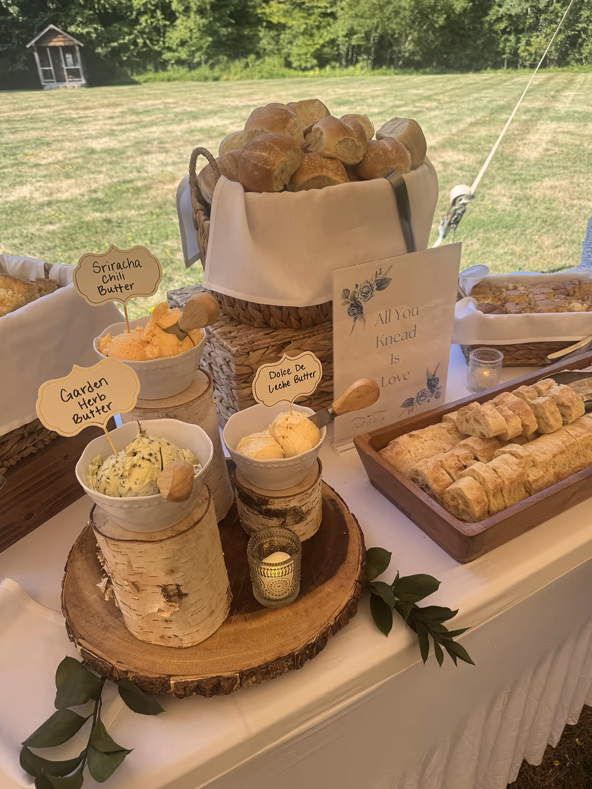 Table with assorted bread rolls, three bowls of flavored butter labeled Sriracha Chili Butter, Garden Herb Butter, and Dolce De Leche Butter, and a sign reading All You Knead Is Love outdoors.