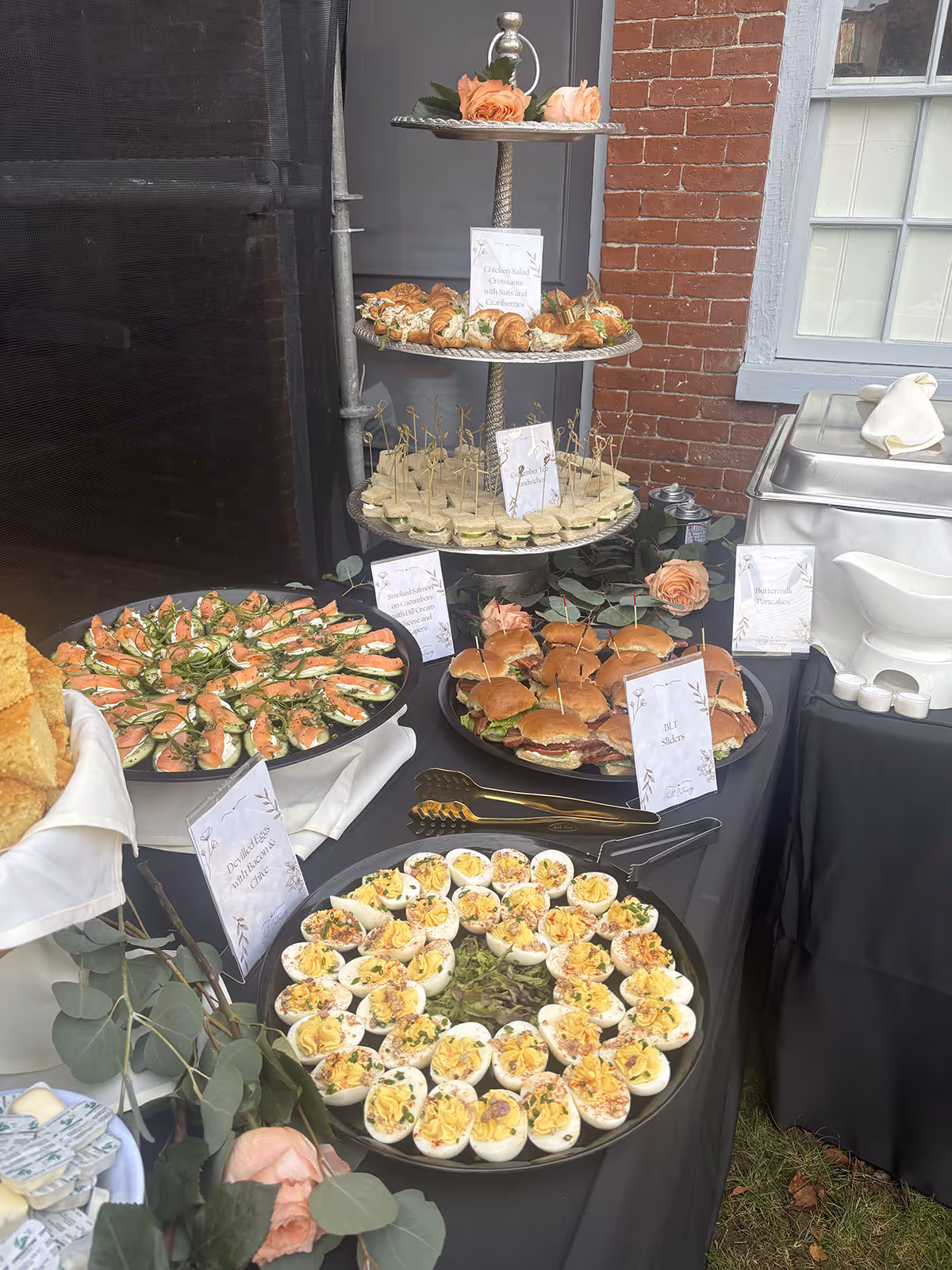 Buffet table with assorted appetizers including deviled eggs, smoked salmon on cucumber slices, BLT sliders, chicken salad croissants, and cucumber tea sandwiches, decorated with pink roses and greenery.