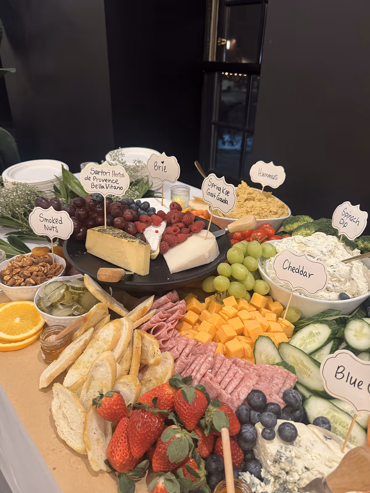Cheese and charcuterie board with labeled items including Brie, Spring Koe Goat Gouda, Sartori Herbs de Provence Bella Vitano, smoked nuts, hummus, spinach dip, cheddar, blue cheese, fresh strawberries, grapes, cucumbers, pickles, orange slices, and assorted bread slices.