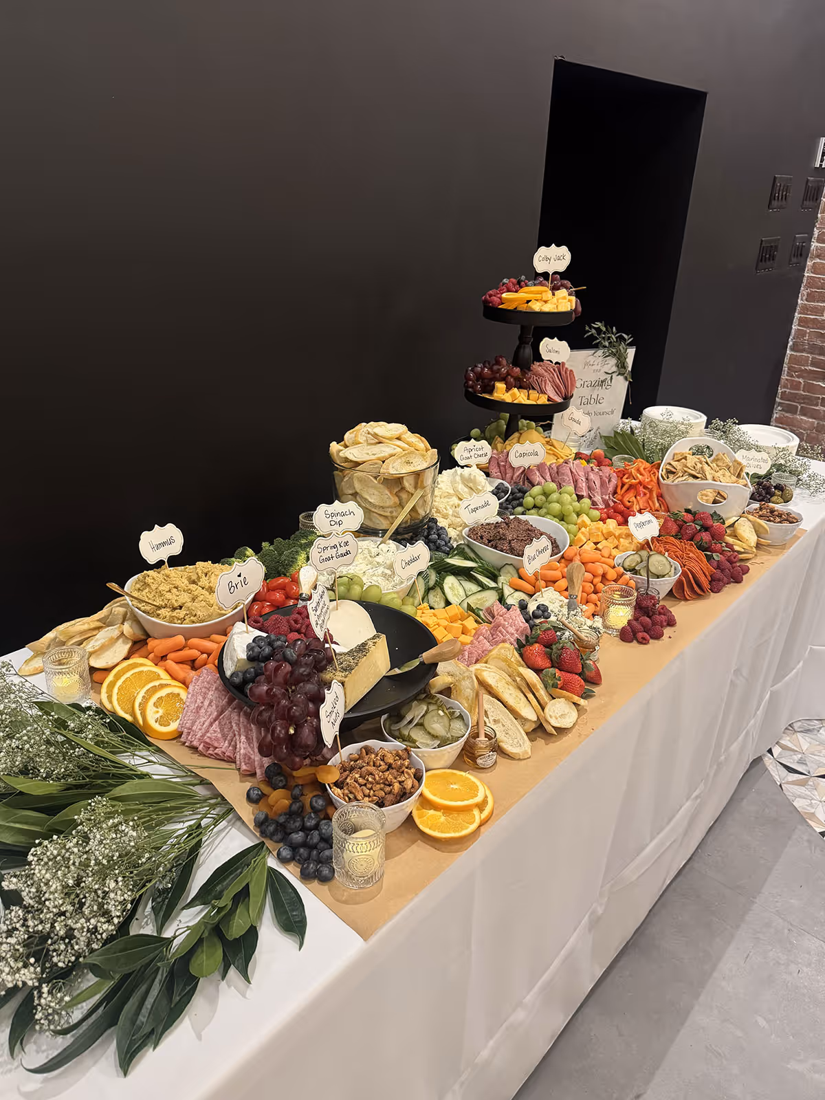 A large grazing table with assorted cheeses, meats, crackers, pickles, fruits, nuts, and dips, arranged on a table with white tablecloth and greenery decorations.