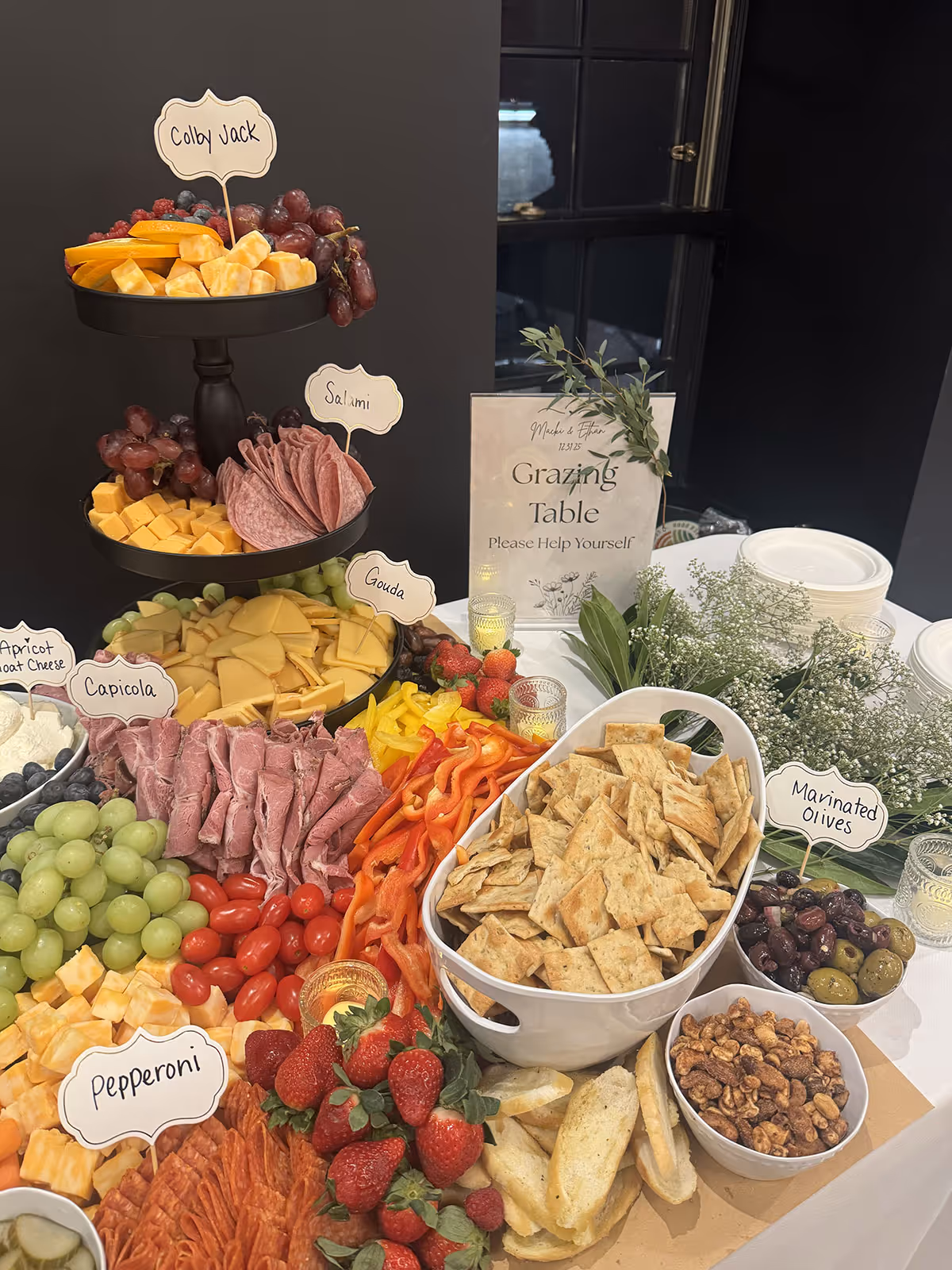 Grazing table with tiered trays of Colby Jack cheese, salami, Gouda, and Capicola, surrounded by grapes, strawberries, sliced peppers, crackers, marinated olives, nuts, and apricot goat cheese.