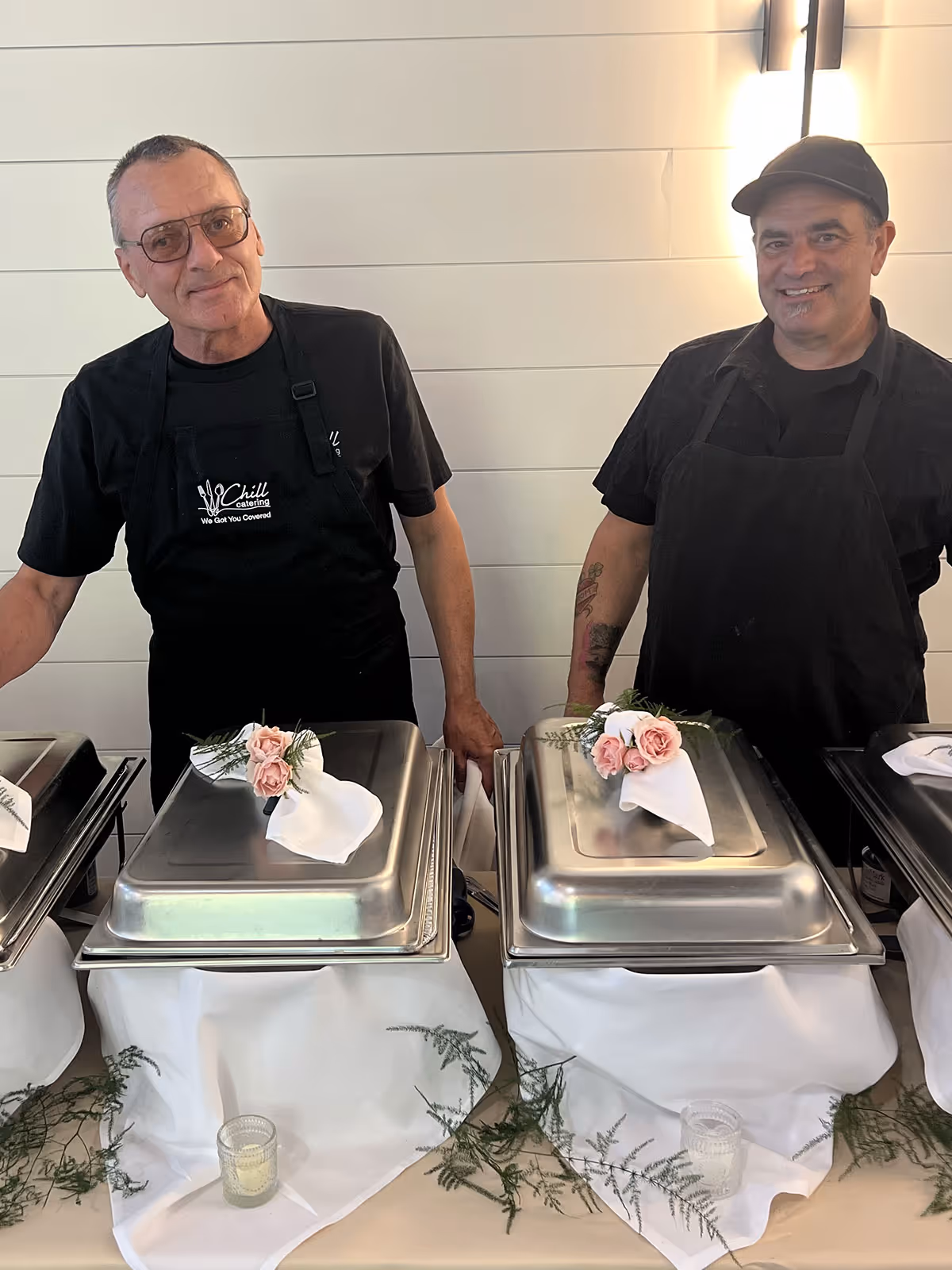 Two male caterers wearing black aprons standing behind buffet chafing dishes decorated with white napkins and pink flowers.