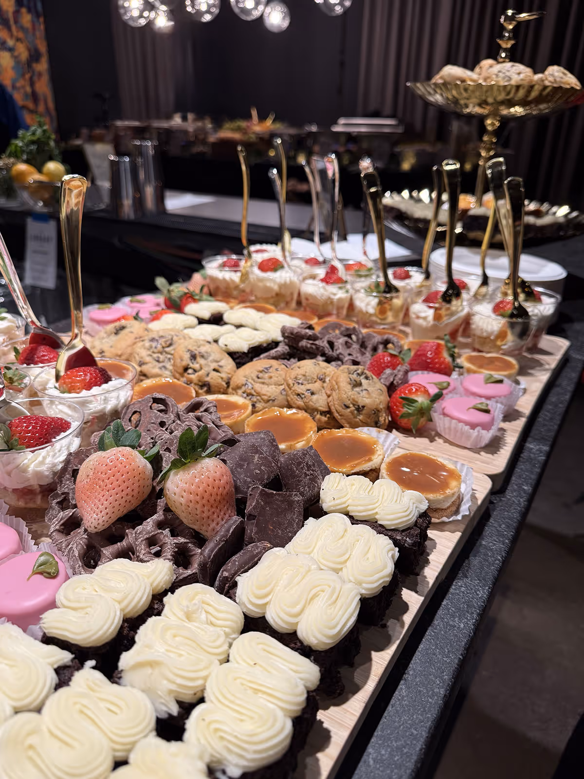 Variety of desserts including cupcakes with cream frosting, chocolate-covered pretzels, cookies, caramel tarts, pink frosted treats, and strawberries arranged on a wooden serving board.