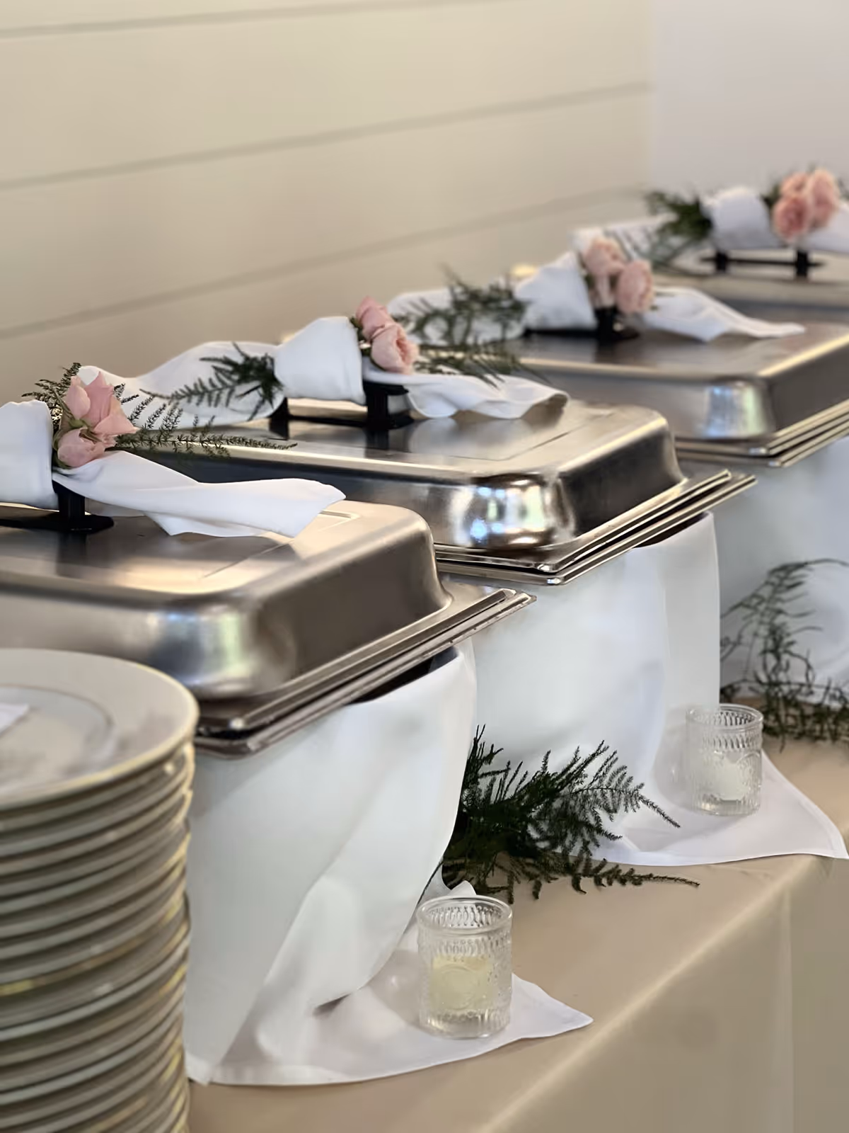 Row of silver chafing dishes decorated with white napkins and pink flowers on a cream-colored tablecloth.