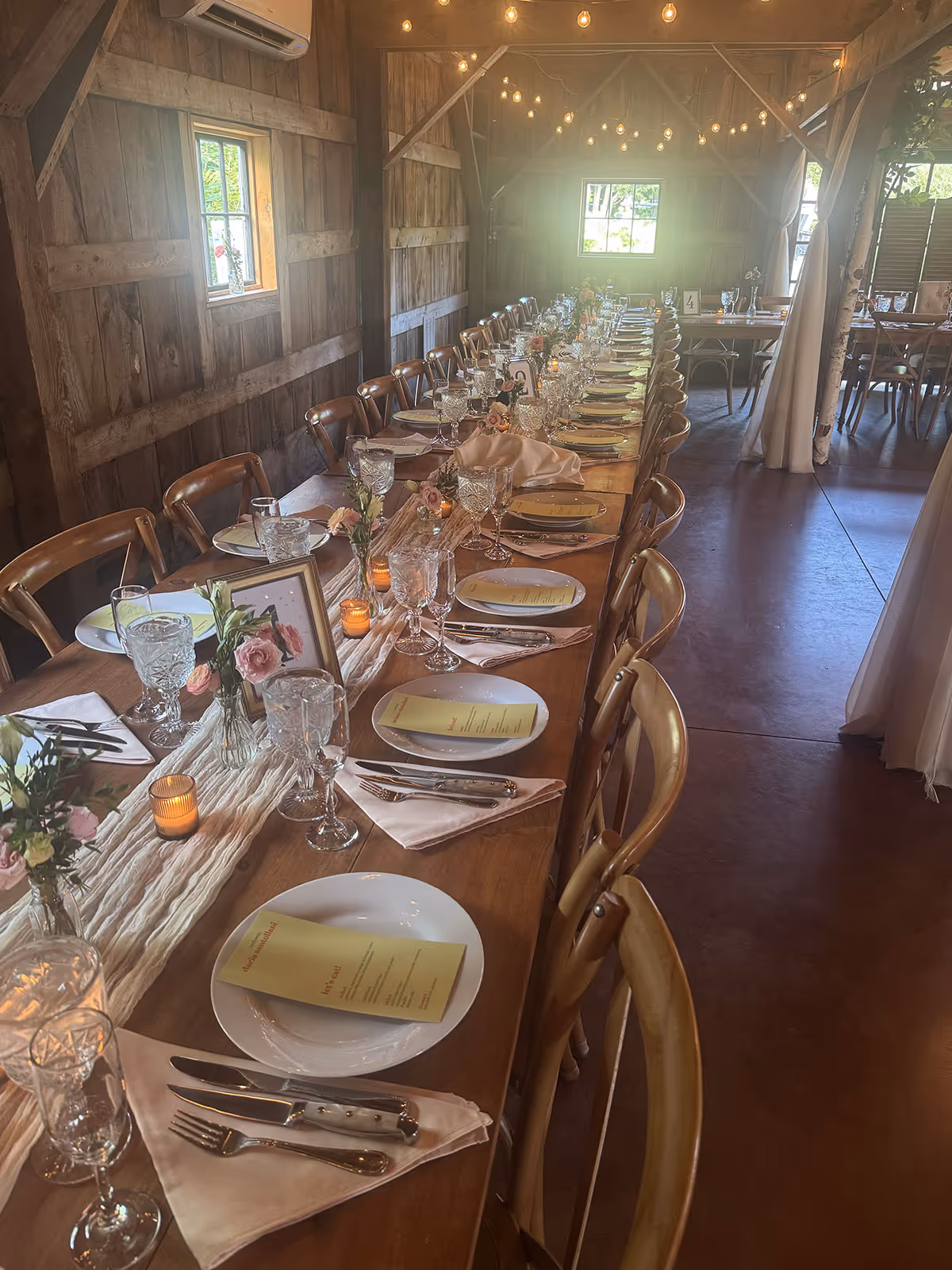 Long wooden dining table set with plates, crystal glasses, silverware, menus, small flower vases, and lit candles in a rustic barn interior.