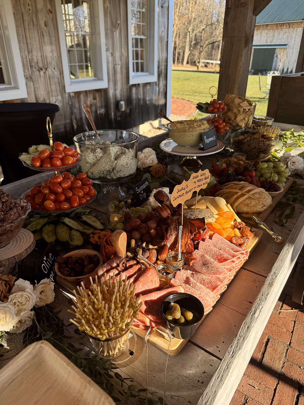 Charcuterie board with assorted sliced meats, cheeses, dried fruits, grapes, pickles, dips, cherry tomatoes, and bread displayed on a wooden table outdoors.