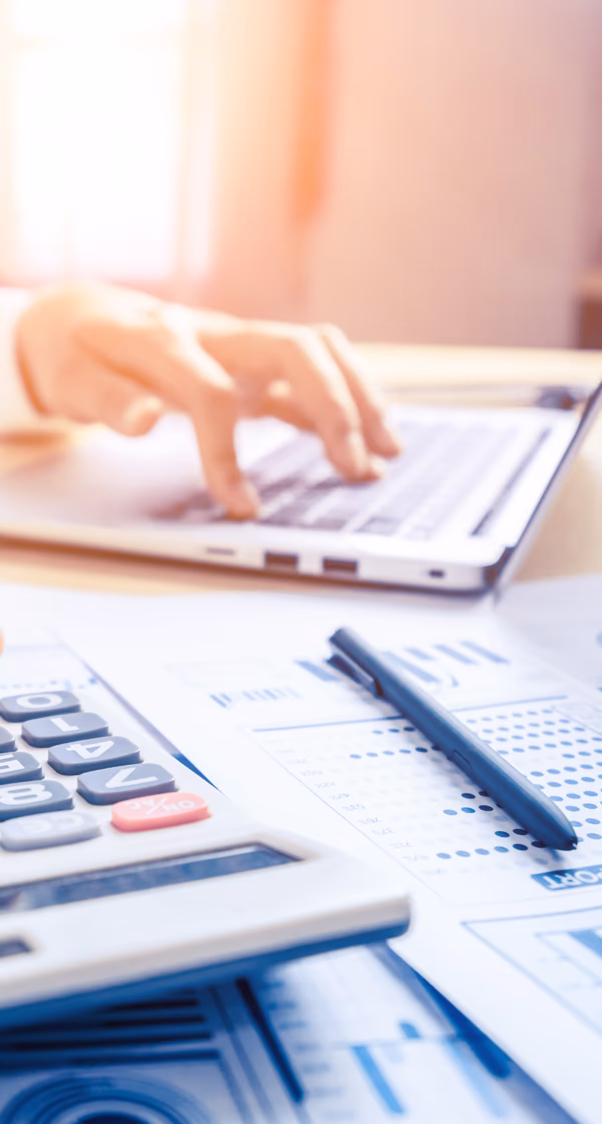 Close-up of a person working on a laptop with financial documents, a pen, and a calculator spread out on a desk. The image suggests accounting, budgeting, or business analysis in a bright office setting.
