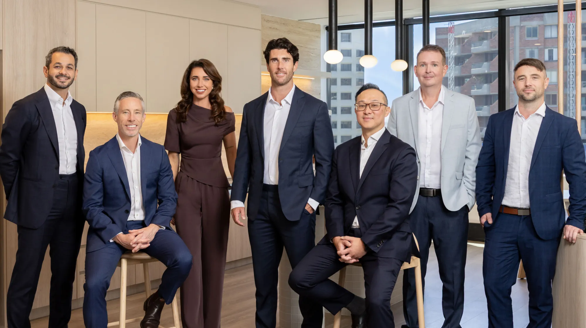 Seven professionally dressed individuals posing in a modern office space with large windows and pendant lights, five standing and two seated on stools.