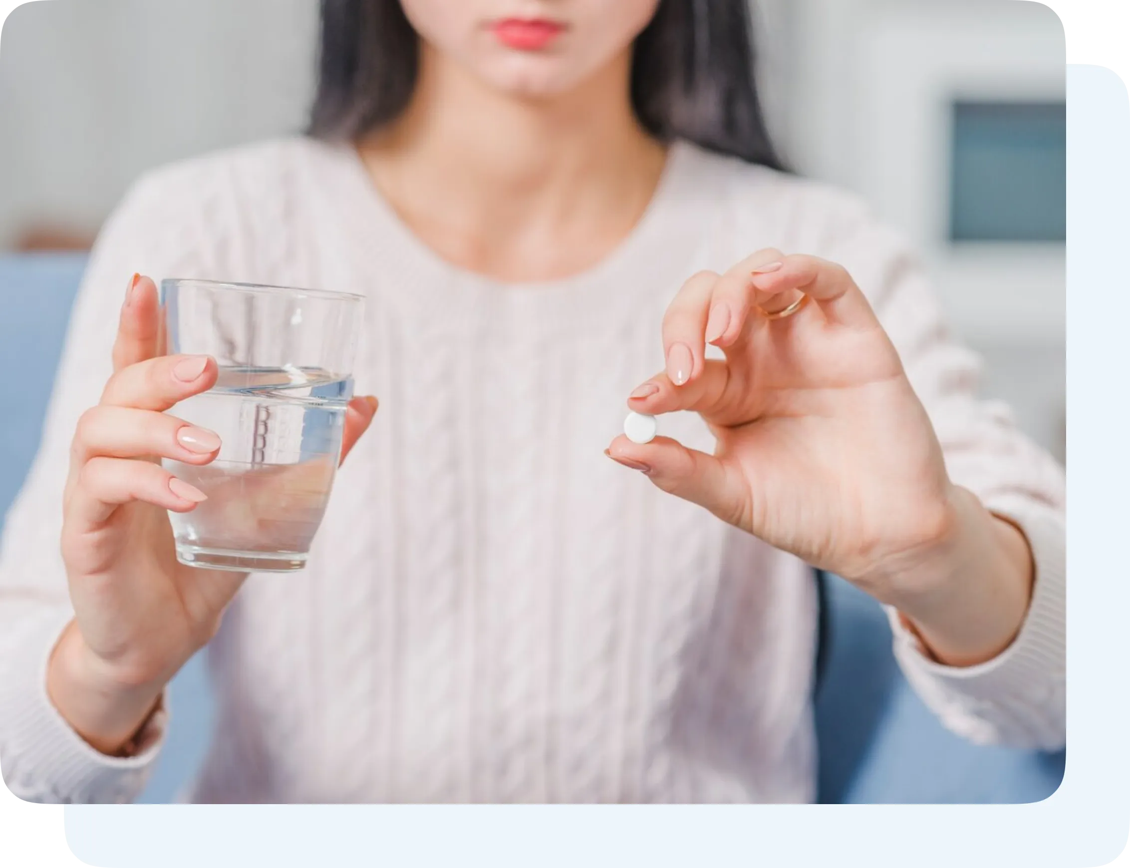 Woman holding a glass of water in one hand and a white pill between fingers of the other hand.