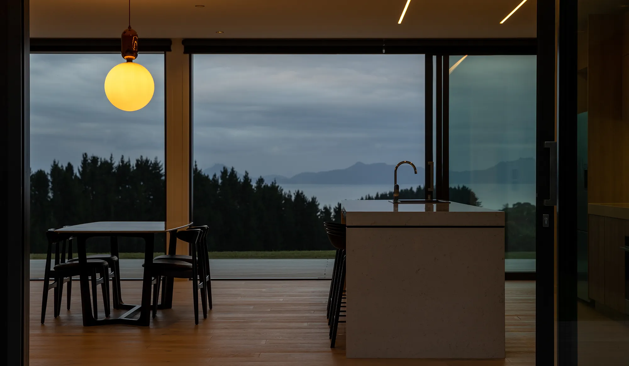 Modern kitchen interior with a large island and dining table, overlooking a forest and distant mountains through floor-to-ceiling windows at dusk.