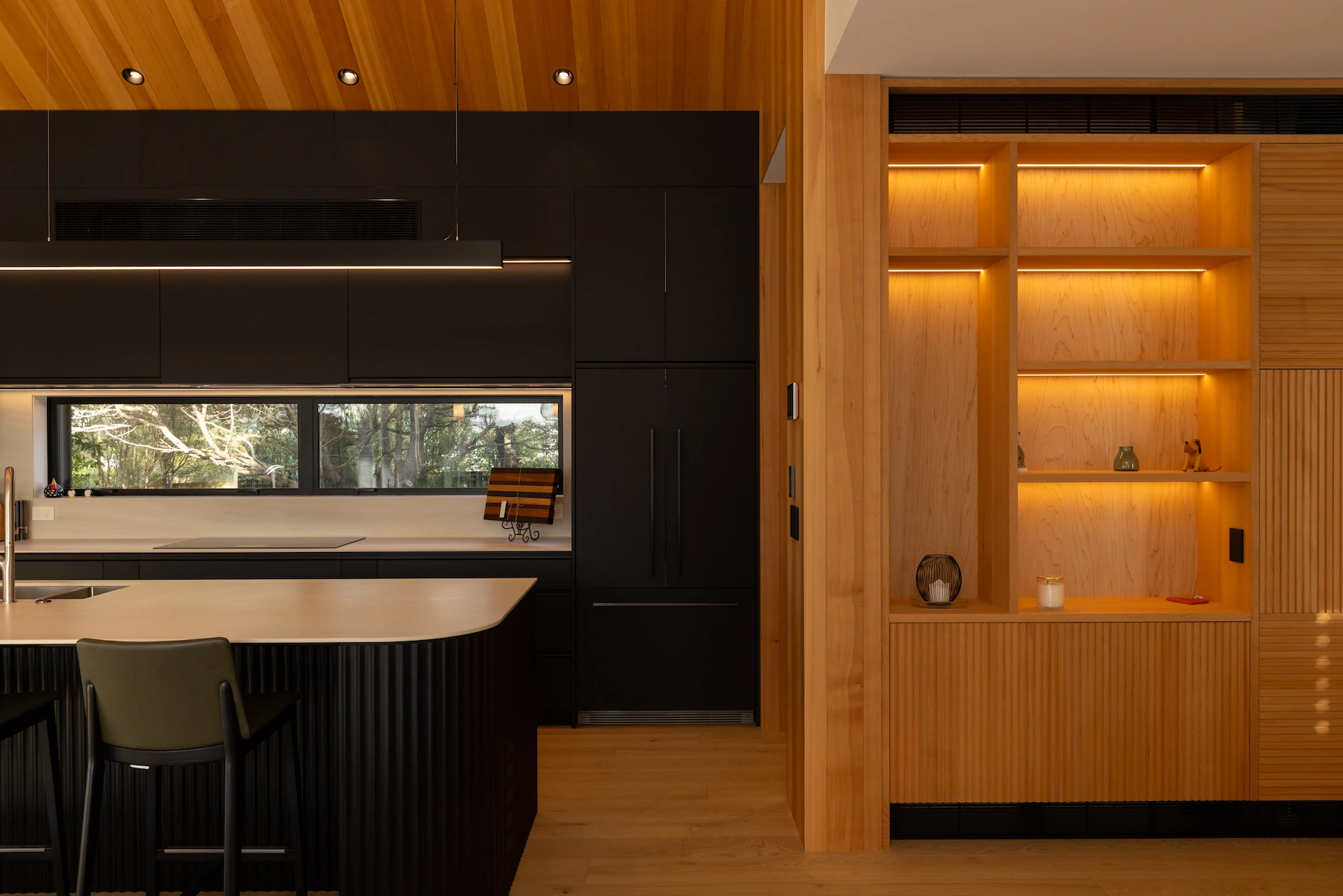 Modern kitchen with black cabinets, beige island, wooden ceiling, and lit wooden shelving unit.