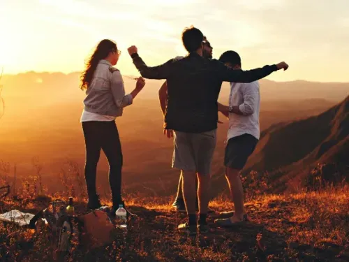 Group of friends enjoying a sunset on a mountaintop, standing together and celebrating, with scenic views of mountains and a warm golden sky in the background.