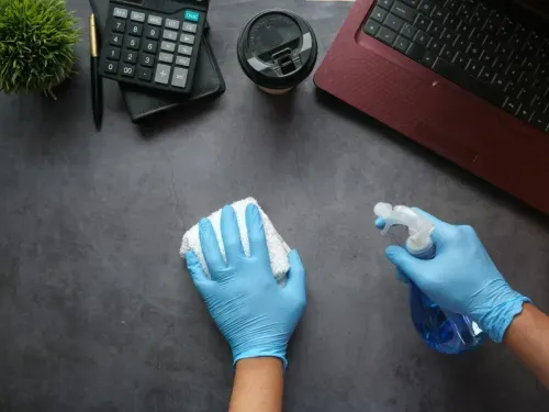 Person cleaning a desk with a spray bottle and cloth, wearing blue gloves, next to a calculator, coffee cup, and laptop.