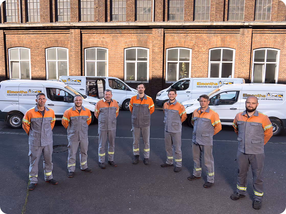 Sieben Mitarbeiter in Arbeitskleidung stehen vor einem Backsteingebäude mit Firmenfahrzeugen von Elmontha im Hintergrund.