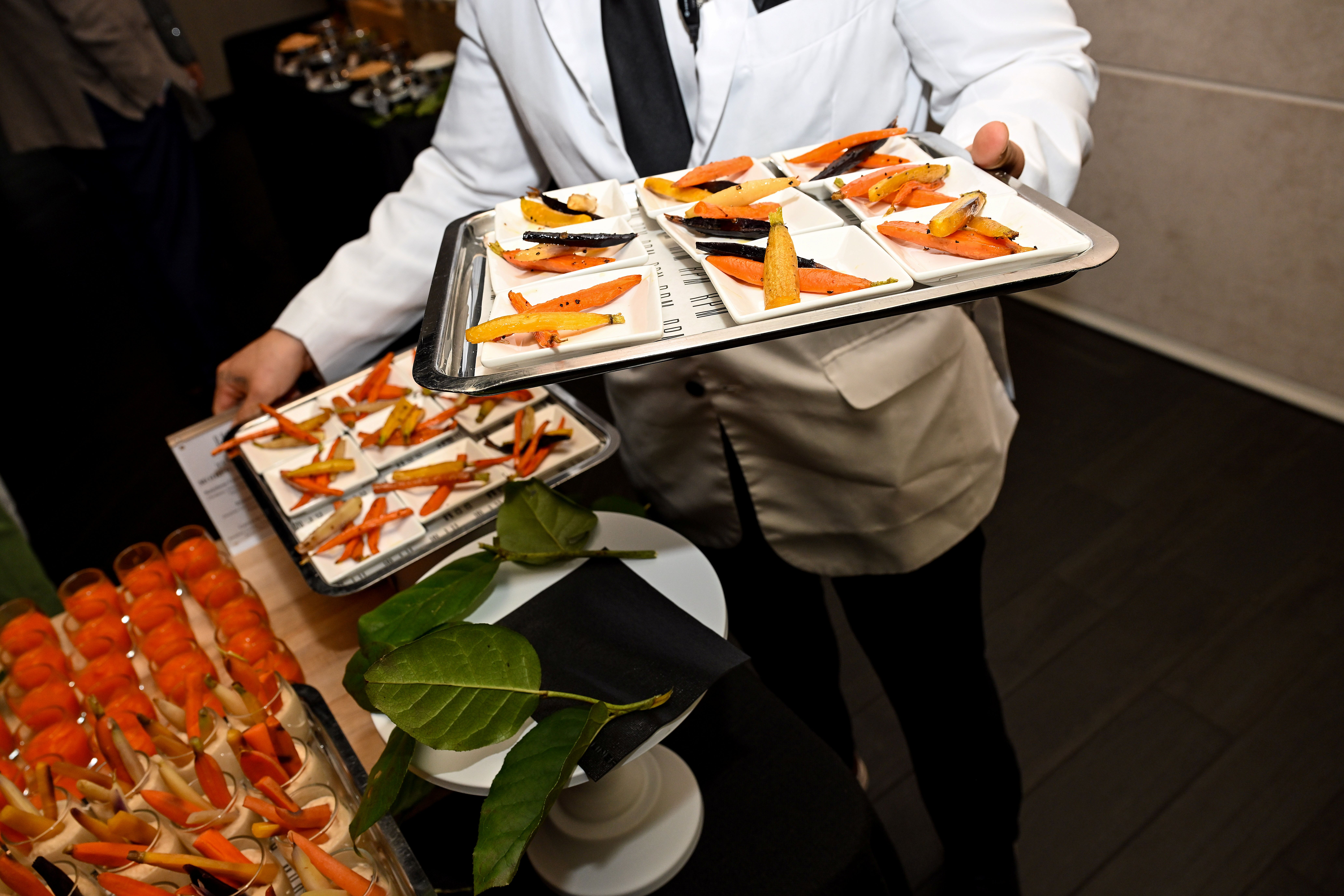 Waiter in white jacket holding trays with small plates of assorted carrot sticks at event.