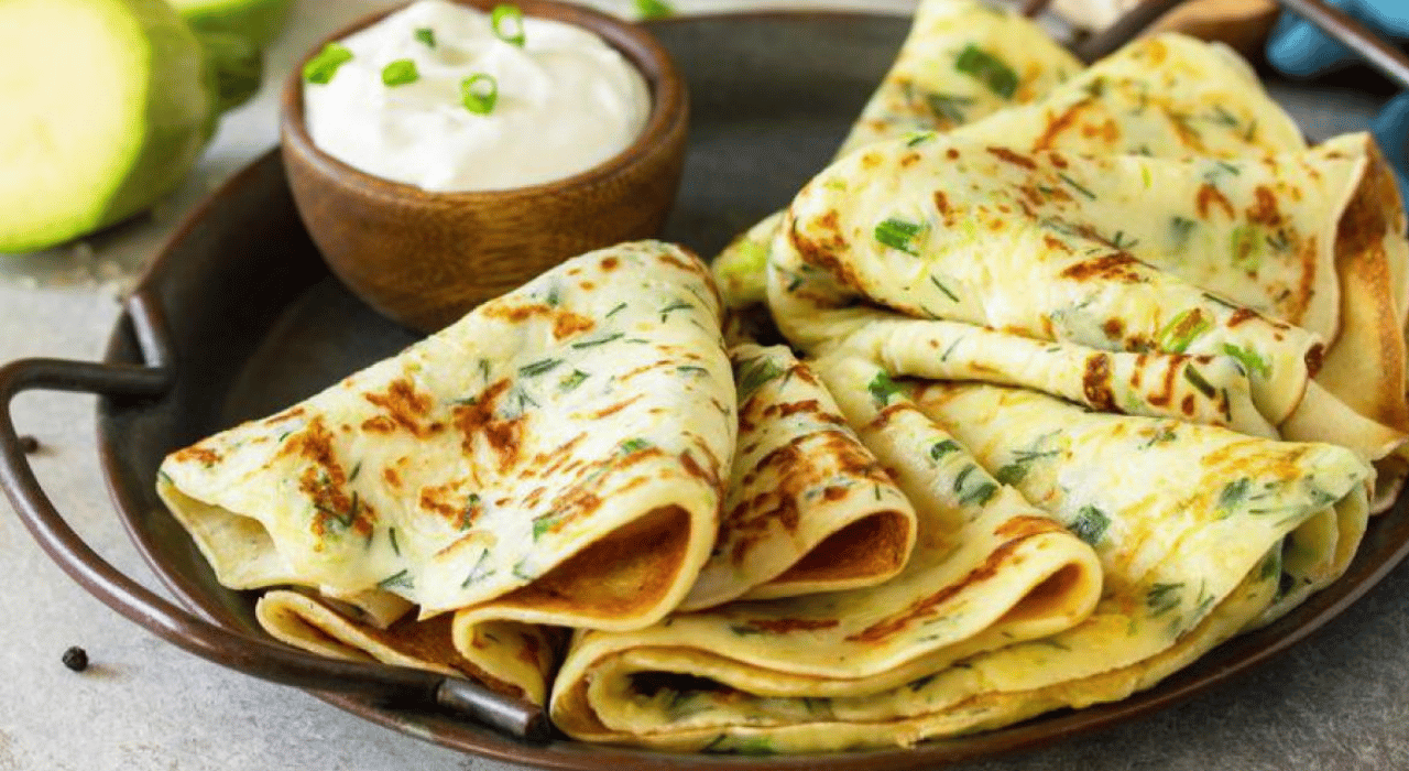 Several zucchini crepes folded over artfully displayed on a plate with a small bowl of yogurt on the side