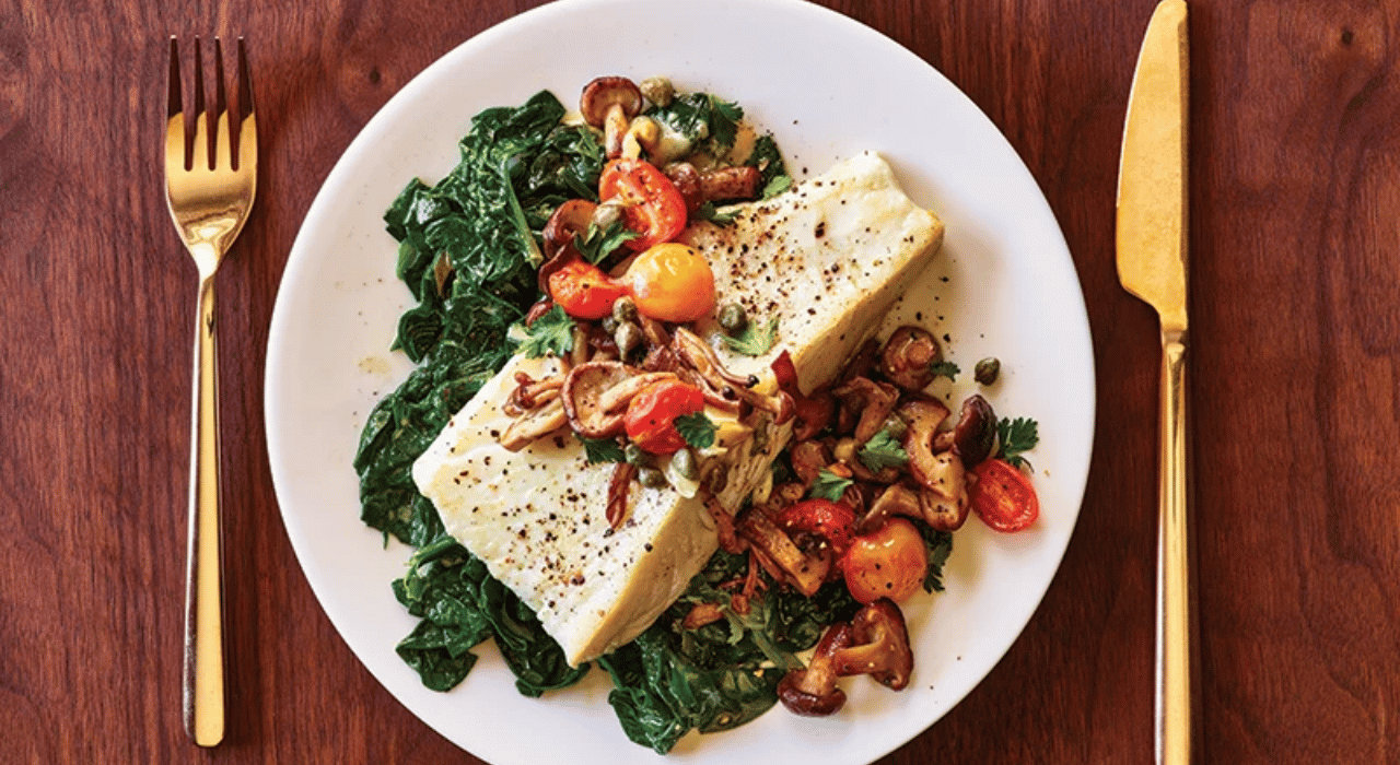 White wine halibut served on a plate next to sauteed spinach and topped with sauteed tomatoes and mushrooms.