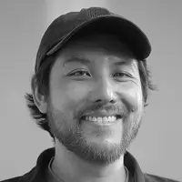 Smiling man with a beard wearing a black baseball cap and dark shirt against a gray background.