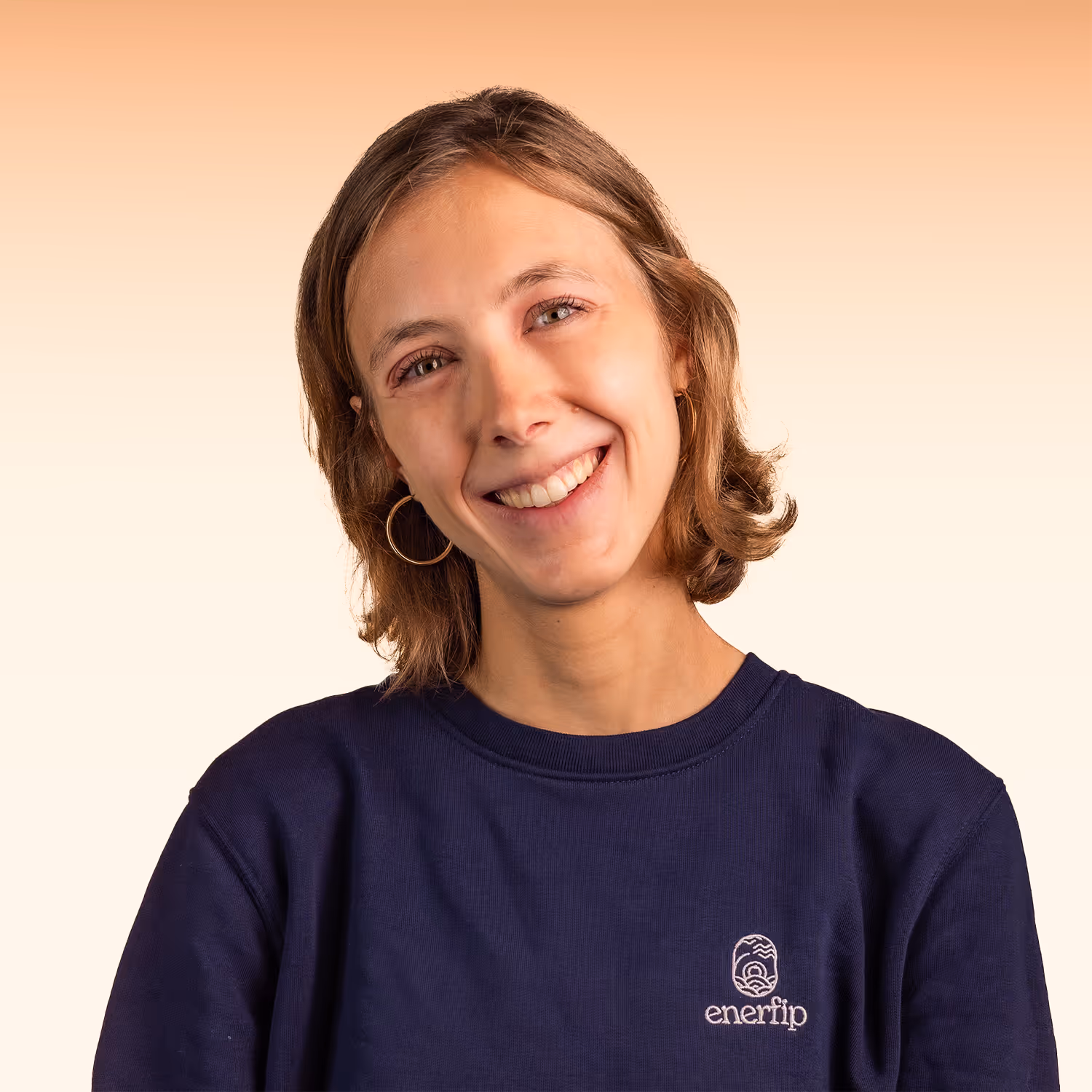 Jonge vrouw met kort bruin haar en gouden oorringen glimlachend in een donkerblauw Enerfip-trui voor een beige achtergrond.