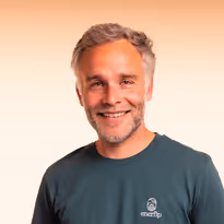 Lachende man met lichtgrijs haar en baard draagt een donkergroene trui met een logo tegen een lichte achtergrond.