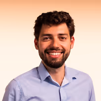 Een glimlachende man met kort krullend haar en een lichte baard, gekleed in een lichtblauw overhemd tegen een zachte beige achtergrond.