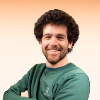 Een man met donkere, krullende haar en baard draagt een groen sweatshirt en lacht met gekruiste armen tegen een peachkleurige achtergrond.