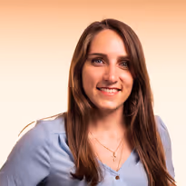 Vrouw met lang bruin haar en blauwe blouse glimlachend tegen een lichte achtergrond.