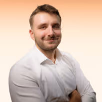 Een man met blond haar en lichte baard die glimlacht, gekleed in een wit overhemd met gekruiste armen tegen een zachte oranje achtergrond.