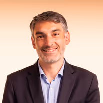 Een glimlachende man met kort grijs haar draagt een donker colbert en een lichtblauw overhemd tegen een effen beige achtergrond.