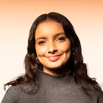 Vrouw met lang, donker haar en een grijze trui glimlachend tegen een lichte achtergrond.