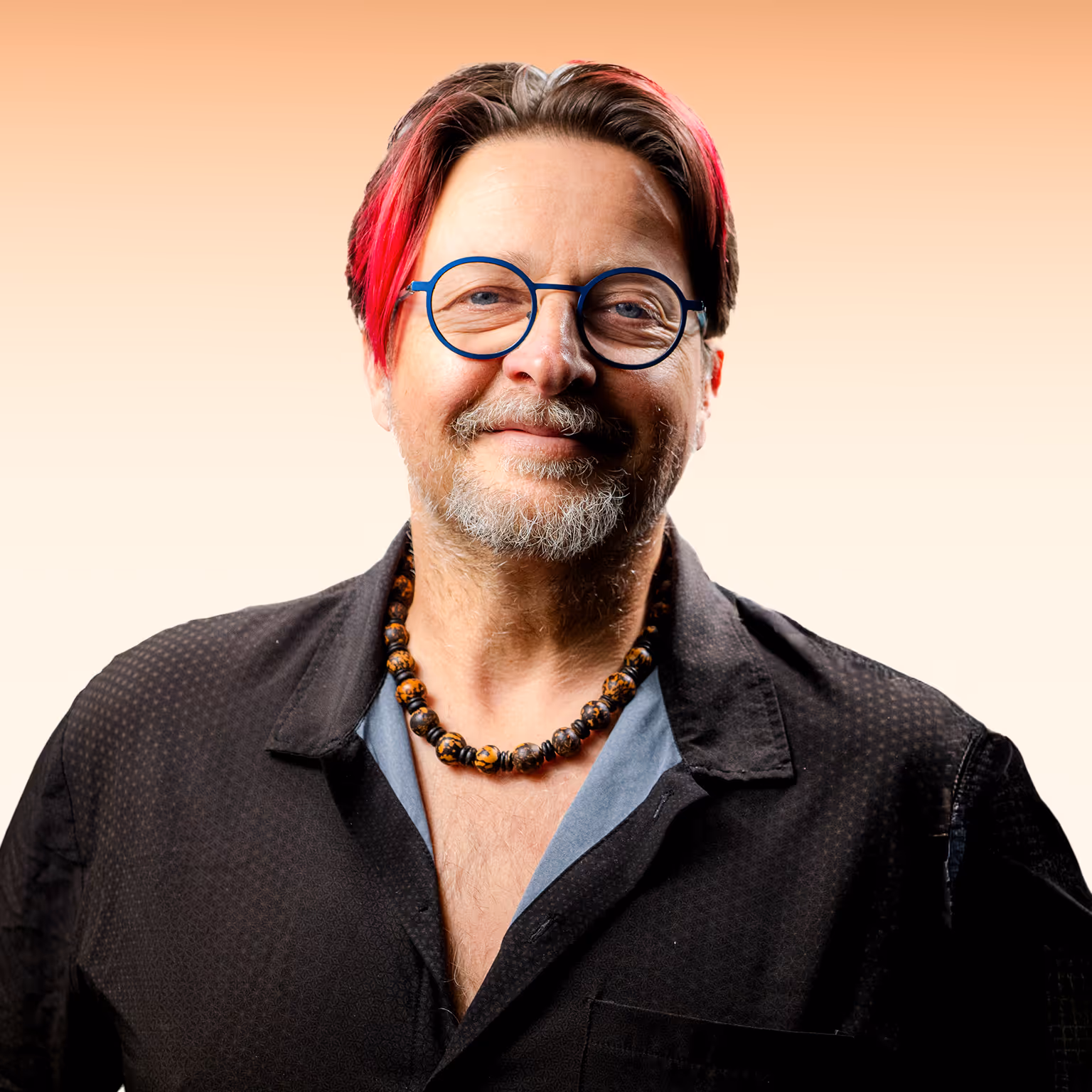 Man met rood geverfd haar aan de zijkanten, blauwe ronde bril en kralenketting, glimlachend tegen een lichte achtergrond.