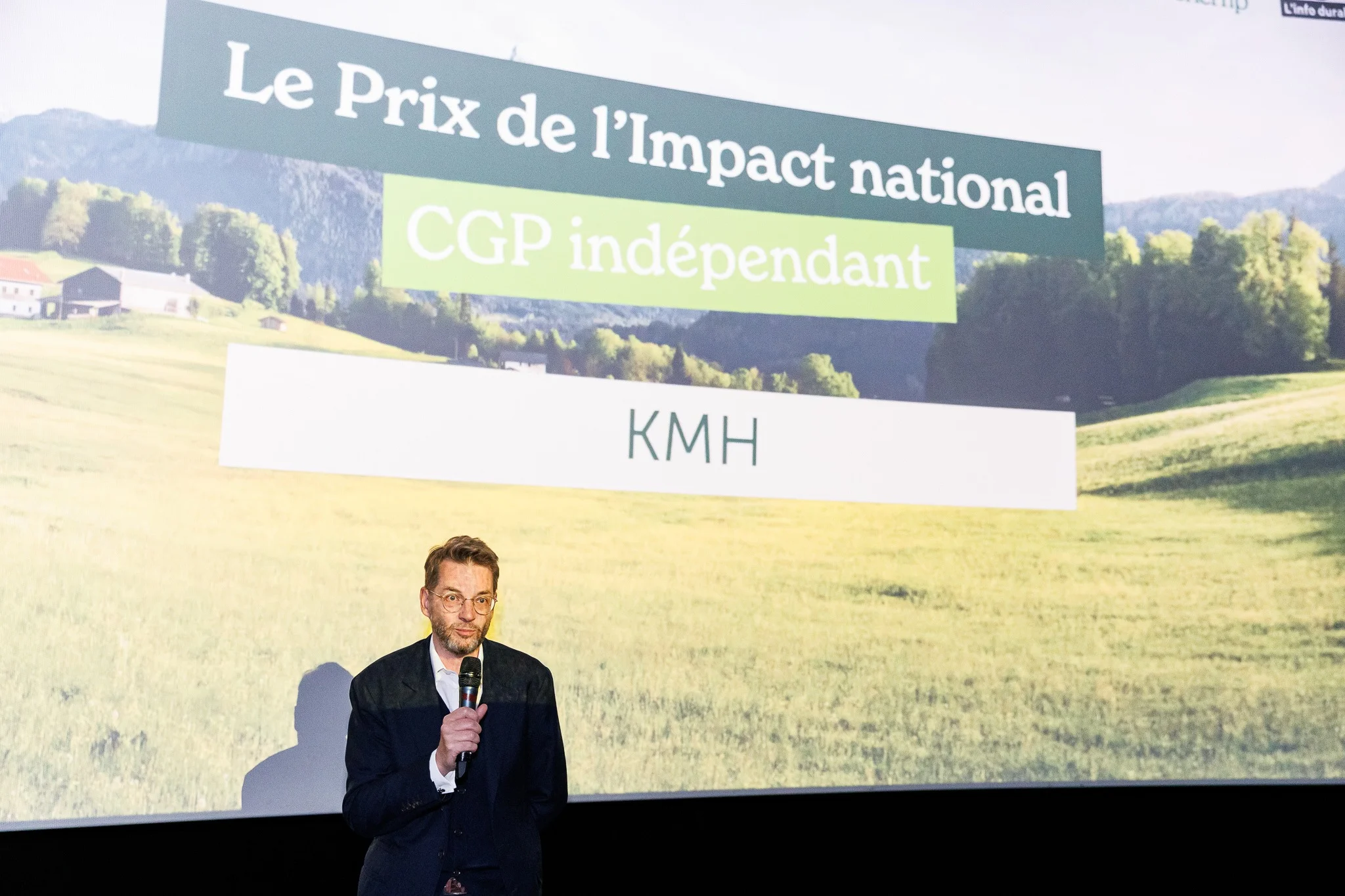 Un homme muni d'un micro, debout devant un écran sur lequel est inscrit « Le Prix de l'Impact national CGP indépendant KMH ».