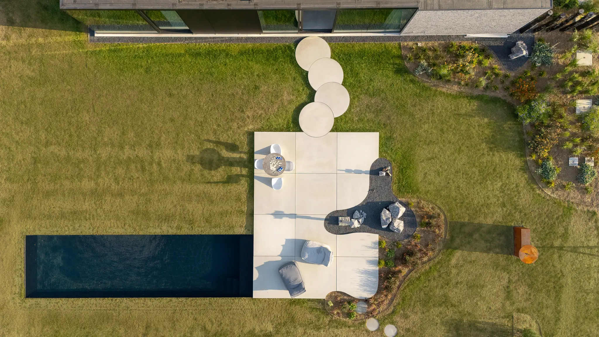 In deze tuin, met adembenemend uitzicht op de omliggende velden, draait alles om natuurlijke overgangen en openheid, de tuin loopt naadloos over in het omliggende landschap, zonder rechte lijnen maar met vloeiende vormen.
De ronde vloerplaten, in de vorm van een wassende maan, creëren een perfecte verbinding tussen de moderne woning en het zonneterras met het aangrenzende zwembad.
In dit minimalistische ontwerp is gekozen om de ruimte maximaal te laten aansluiten op de omgeving, waardoor elke hoek uitnodigt tot ontspanning en genot van de natuur. Kortom: een harmonieus samenspel tussen architectuur en natuur