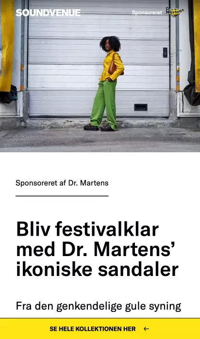 Person in yellow jacket, green pants, and Dr. Martens sandals standing in front of a closed industrial garage door.