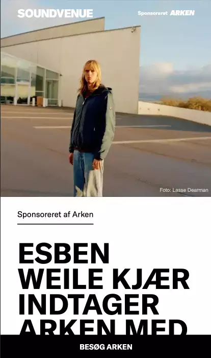 Blonde person wearing a blue jacket and jeans standing outdoors near a modern building with clear sky backdrop.