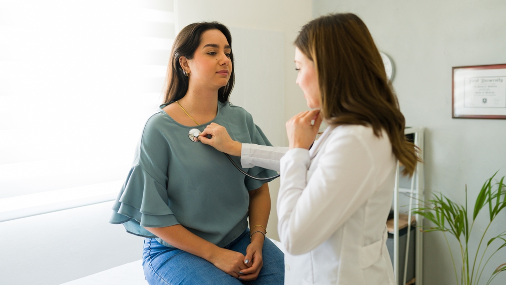 Doctor is listening to a female patient's heart while holding a stethoscope to her chest in a medical office